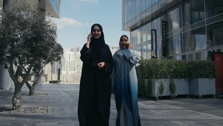 Two Saudi Arabian Gulf women walking together talking on the mobile phone, two smiling Emirati friends walking together on a pleasant outing to a recreational place, tourism and recreation, friendships and acquaintances, social relations, spending a pleasant time, talking with friends