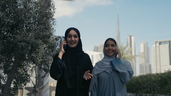 سيدتان عربيتان خليجيتان سعوديتان تسيران سويا تتحدثان عبر الهاتف المحمول،  صديقتان اماراتيتان مبتسمتان تسيران معاً في نزهة ممتعة لإحدى الأماكن الترفيهية، سياحة و استجمام، صداقات و تعارف، علاقات إجتماعية، قضاء أوقات ممتعة، التحدث برفقه الاصدقاء