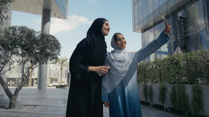 توثيق اللحظات السعيدة، استخدام الأجهزة والتقنيات المتطورة، امرأتان عربيتان خليجيتان محجبتان ترتديان العباءة الخليجيه تقومان بالتقاط صور سيلفي تذكارية بإيماءات السعادة والسرور، سيدتان سعوديتان تقومان بالتقاط صور تذكاريه