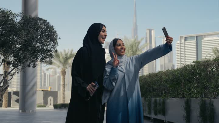 توثيق اللحظات السعيدة، استخدام الأجهزة والتقنيات المتطورة، امرأتان عربيتان خليجيتان محجبتان ترتديان العباءة الخليجيه تقومان بالتقاط صور سيلفي تذكارية بإيماءات السعادة والسرور، سيدتان سعوديتان تقومان بالتقاط صور تذكاريه