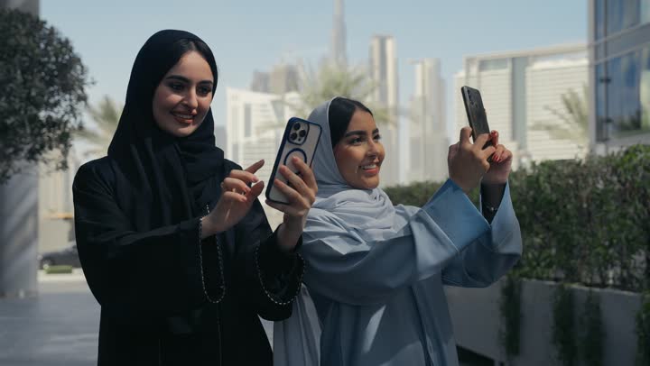 Two smiling Saudi Arabian Gulf Arab women walking together on a pleasant outing to an entertainment venue, two Emirati friends taking a souvenir selfie with their mobile phones, friendships and acquaintances, social relations
