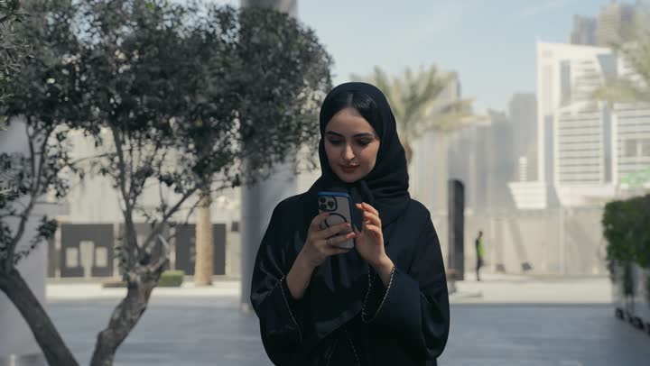 A Saudi Arabian Gulf woman using a mobile phone, gestures and facial expressions indicating happiness, exchanging news via text messages, chatting with family and friends, an Emirati woman messaging online, using modern technologies