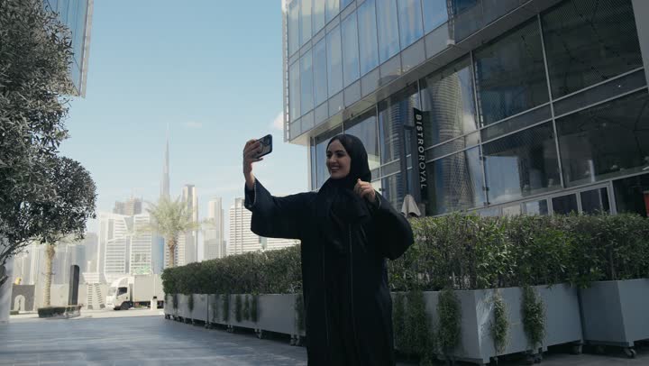 Documenting happy moments, using modern technical devices, a smiling veiled Saudi Arabian Gulf woman holding a mobile phone in her hand, taking souvenir selfies, various mobile programs and applications, an Emirati woman wearing a colorful abaya walking in the city