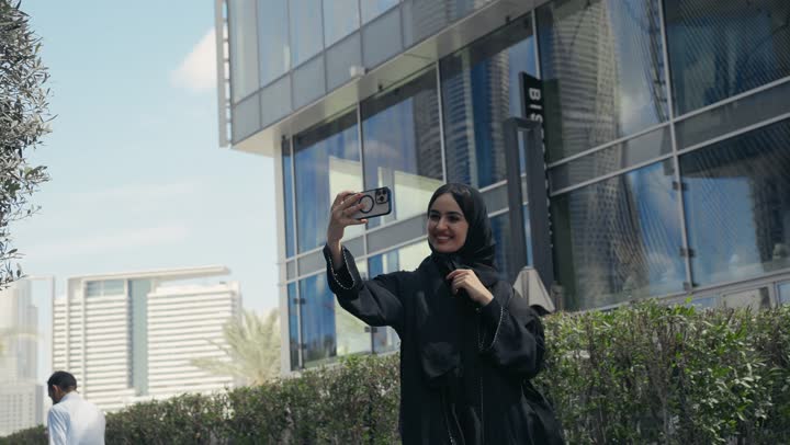 توثيق اللحظات السعيدة، إستخدام الأجهزة التقنية الحديثة، امرأه عربية خليجية سعودية محجبة مبتسمة تمسك بيدها جهاز الهاتف المحمول تقوم بالتقاط صور السيلفي التذكارية، برامج وتطبيقات الجوال المتنوعة، سيده اماراتيه ترتدي عباءه ملونه تسيرفي المدينه