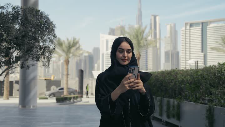 امرأه عربية خليجية سعودية محجبة مبتسمة تمسك بيدها جهاز الهاتف المحمول تقوم بتصوير فيديو لاحد الاماكن الترفيهيه، سيده سعوديه ترتدي عباءه سوداء تستخدم جهاز تقني حديث، توثيق اللحظات السعيدة، إستخدام الأجهزة التقنية الحديثة