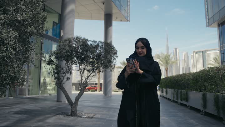 A smiling veiled Saudi Arabian Gulf woman holding a mobile phone in her hand, filming a video of an entertainment venue. A Saudi woman wearing a black abaya using a modern technical device, documenting happy moments, using modern technical devices.