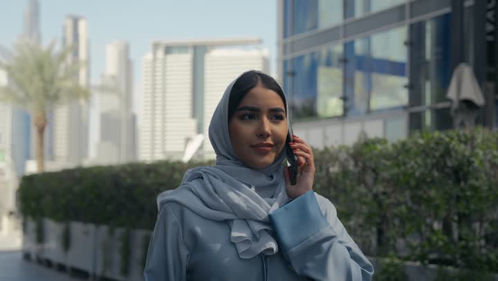 الانزعاج بسماع اخبار سيئه، تبادل الاخبار والأحاديث مع الاصدقاء والأهل، إمرأة عربية سعودية خليجية محجبة ترتدي العباءة تقوم بالتحدث عبر الهاتف المحمول، ايماءات السعادة و السرور بإجراء مكالمة هاتفية، سيده اماراتيه ترتدي عباءه ملونه تسيرفي مكان خارجي