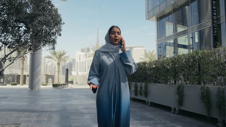 قضاء اوقات ممتعة بتبادل الاخبار والأحاديث مع الاصدقاء والأهل، إمرأة عربية سعودية خليجية محجبة ترتدي العباءة تقوم بالتحدث عبر الهاتف المحمول، ايماءات السعادة و السرور بإجراء مكالمة هاتفية، سيده اماراتيه ترتدي عباءه ملونه تسيرفي مكان خارجي