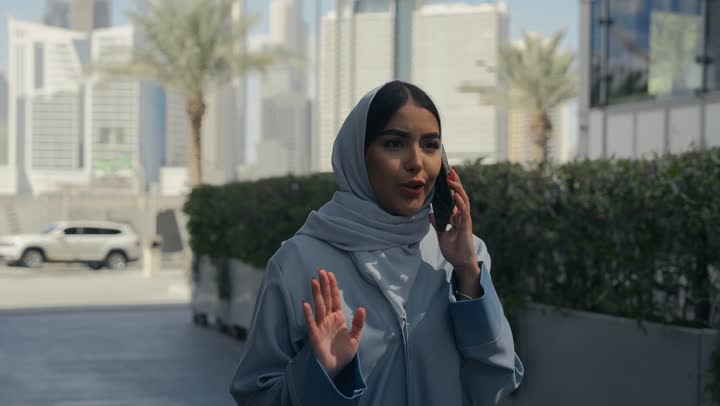 Having a good time exchanging news and conversations with friends and family, a veiled Saudi Arabian Gulf woman wearing an abaya talking on a mobile phone, gestures of happiness and pleasure while making a phone call, an Emirati woman wearing a colorful abaya walking in an outdoor place