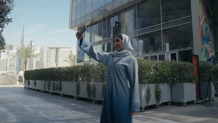 توثيق اللحظات السعيدة، إستخدام الأجهزة التقنية الحديثة، امرأه عربية خليجية سعودية محجبة مبتسمة تمسك بيدها جهاز الهاتف المحمول تقوم بالتقاط صور السيلفي التذكارية، سيده سعوديه ترتدي عباءه رماديه تستخدم جهاز تقني حديث