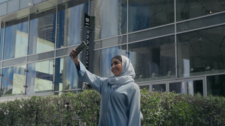 توثيق اللحظات السعيدة، إستخدام الأجهزة التقنية الحديثة، امرأه عربية خليجية سعودية محجبة مبتسمة تمسك بيدها جهاز الهاتف المحمول تقوم بالتقاط صور السيلفي التذكارية، سيده سعوديه ترتدي عباءه رماديه تستخدم جهاز تقني حديث