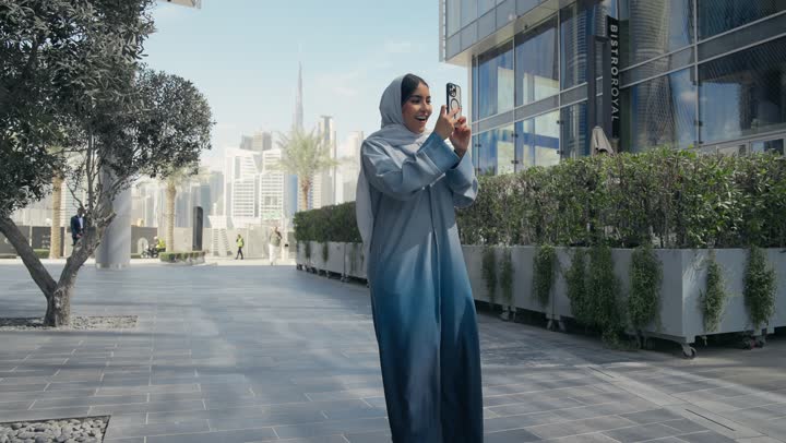 برامج وتطبيقات الجوال المتنوعة، توثيق اللحظات السعيدة، إستخدام الأجهزة التقنية الحديثة، امرأه عربية خليجية سعودية محجبة مبتسمة تمسك بيدها جهاز الهاتف المحمول تقوم بالتقاط صور السيلفي التذكارية، سيده سعوديه ترتدي عباءه رماديه تستخدم جهاز تقني حديث