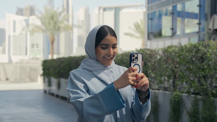 برامج وتطبيقات الجوال المتنوعة، توثيق اللحظات السعيدة، إستخدام الأجهزة التقنية الحديثة، امرأه عربية خليجية سعودية محجبة مبتسمة تمسك بيدها جهاز الهاتف المحمول تقوم بالتقاط صور السيلفي التذكارية، سيده سعوديه ترتدي عباءه رماديه تستخدم جهاز تقني حديث