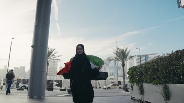 ايماءات وجه تدل علي الشموخ والافتخار، يوم الاستقلال الوطني الاماراتي،  سيده اماراتيه تحمل علم دولة الامارات علي ظهرها، ذكرى اليوم الوطني الاماراتي، امرأه اماراتية ترتدي عباءه سوداء تحتفل باليوم الوطني لدولة الامارات العربية المتحدة