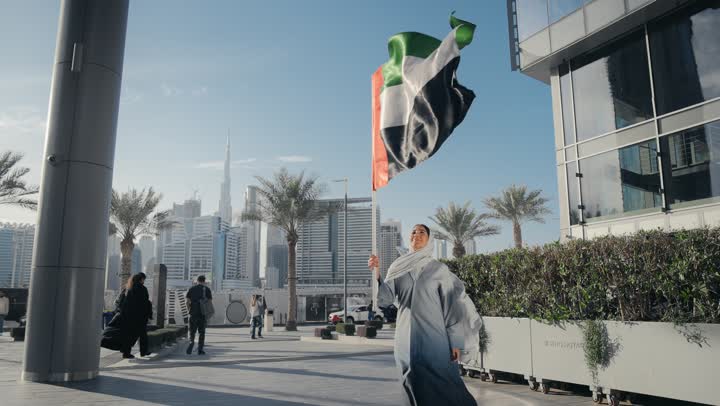 سيده اماراتيه تحمل بيدها علم دولة الامارات وتقوم برفعه الي الاعلي، ايماءات وجه تدل علي الحب و السعادة، ذكرى اليوم الوطني الاماراتي، امرأه اماراتية ترتدي عباءه رماديه تحتفل باليوم الوطني لدولة الامارات العربية المتحدة، يوم الاستقلال الوطني الاماراتي