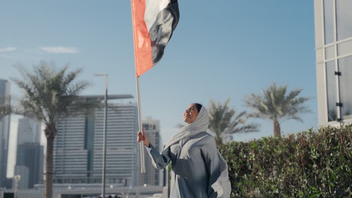Emirati woman holding the UAE flag and raising it high, facial expressions indicating love and happiness, UAE National Day, Emirati woman wearing a gray abaya celebrating UAE National Day, UAE National Independence Day