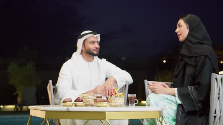 شرب القهوة العربية، قضاء أوقات عائلية ممتعة, التفاهم والترابط الفكري بين الزوجين، تناول الوجبات الخفيفة والتسالي، الجلوس في إحدى الشاليهات الاماراتية الراقية، رجل عربي خليجي اماراتي يرتدي الكندورة والغترة يقوم بتبادل الأحاديث مع زوجته في الهواء الطلق