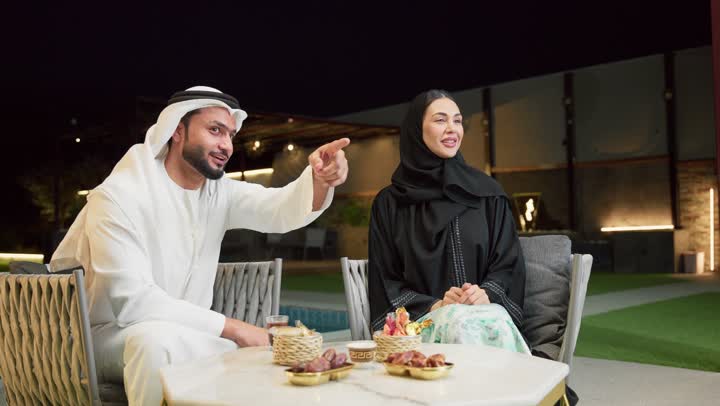 الإشارة إلى شيء ما، قضاء أوقات عائلية ممتعة, التفاهم والترابط الفكري بين الزوجين، تناول الوجبات الخفيفة والتسالي، الجلوس في إحدى الشاليهات الاماراتية الراقية، رجل عربي خليجي اماراتي يرتدي الكندورة والغترة يقوم بتبادل الأحاديث مع زوجته في الهواء الطلق