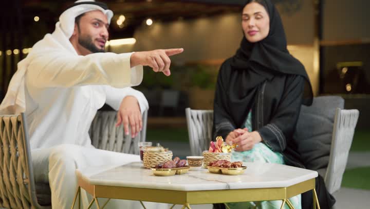 الإشارة إلى شيء ما، قضاء أوقات عائلية ممتعة, التفاهم والترابط الفكري بين الزوجين، تناول الوجبات الخفيفة والتسالي، الجلوس في إحدى الشاليهات الاماراتية الراقية، رجل عربي خليجي اماراتي يرتدي الكندورة والغترة يقوم بتبادل الأحاديث مع زوجته في الهواء الطلق