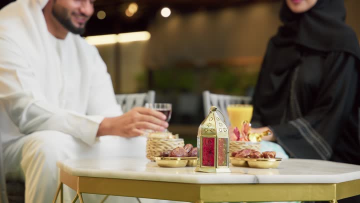 Drinking juices and cold drinks, spending enjoyable family time, understanding and intellectual bonding between spouses, eating snacks and entertainment, sitting in one of the upscale Emirati chalets, an Arab Gulf Emirati man wearing a kandura and ghutra exchanging conversations with his wife in the open air