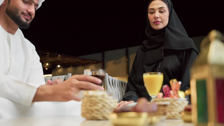 شرب العصائر والمشروبات الباردة، قضاء أوقات عائلية ممتعة, التفاهم والترابط الفكري بين الزوجين، تناول الوجبات الخفيفة والتسالي، الجلوس في إحدى الشاليهات الاماراتية الراقية، رجل عربي خليجي اماراتي يرتدي الكندورة والغترة يقوم بتبادل الأحاديث مع زوجته في الهواء الطلق