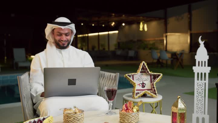 إجراء اجتماع عمل عن بعد عبر الانترنت، جلسة ليلية خارجية في احدى شاليهات الامارات، ضيافة شهر رمضان المبارك، شاب عربي خليجي اماراتي يرتدي الكندورة والغترة يجلس في الهواء الطلق ويقوم باستخدام جهاز الحاسوب بتعابير الاندماج والسرور، زينة واضاءة رمضانية