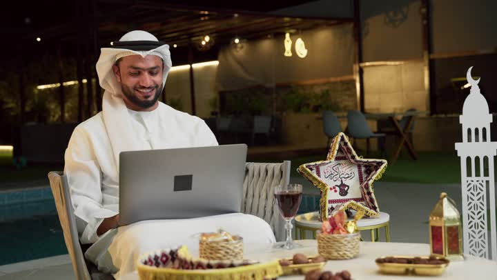 Hosting the holy month of Ramadan, conducting a remote work meeting via the internet, an outdoor evening session in one of the chalets in the Emirates, a young Emirati Gulf Arab man wearing a kandura and ghutra sitting outdoors and using the computer with expressions of integration and pleasure, Ramadan decorations and lighting