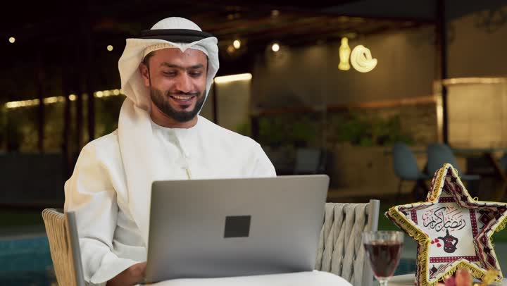Hosting the holy month of Ramadan, conducting a remote work meeting via the internet, an outdoor evening session in one of the chalets in the Emirates, a young Emirati Gulf Arab man wearing a kandura and ghutra sitting outdoors and using the computer with expressions of integration and pleasure, Ramadan decorations and lighting