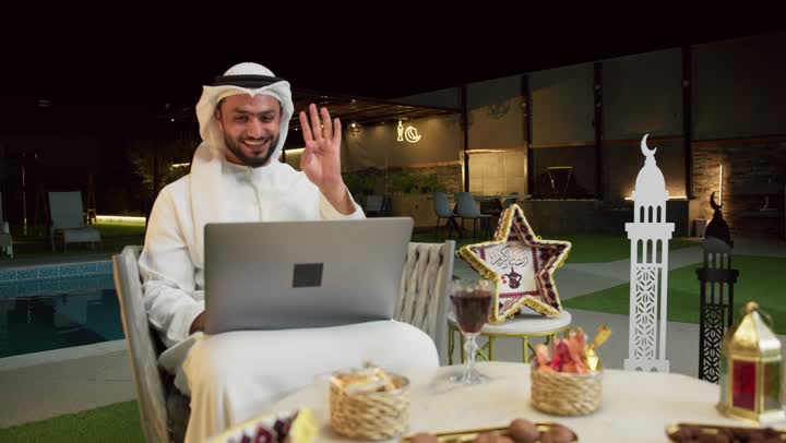 إجراء اجتماع عمل عن بعد عبر الانترنت، جلسة ليلية خارجية في احدى شاليهات الامارات، ضيافة شهر رمضان المبارك، شاب عربي خليجي اماراتي يرتدي الكندورة والغترة يجلس في الهواء الطلق ويقوم باستخدام جهاز الحاسوب بتعابير الاندماج والسرور، زينة واضاءة رمضانية
