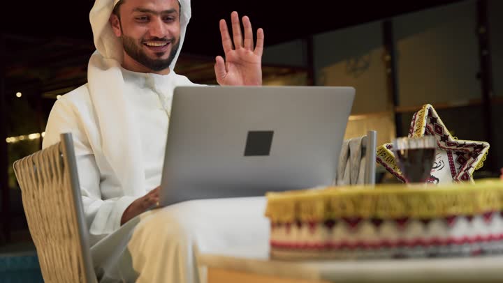 ضيافة شهر رمضان المبارك، إجراء اجتماع عمل عن بعد عبر الانترنت، جلسة ليلية خارجية في احدى شاليهات الامارات، شاب عربي خليجي اماراتي يرتدي الكندورة والغترة يجلس في الهواء الطلق ويقوم باستخدام جهاز الحاسوب بتعابير الاندماج والسرور، زينة واضاءة رمضانية