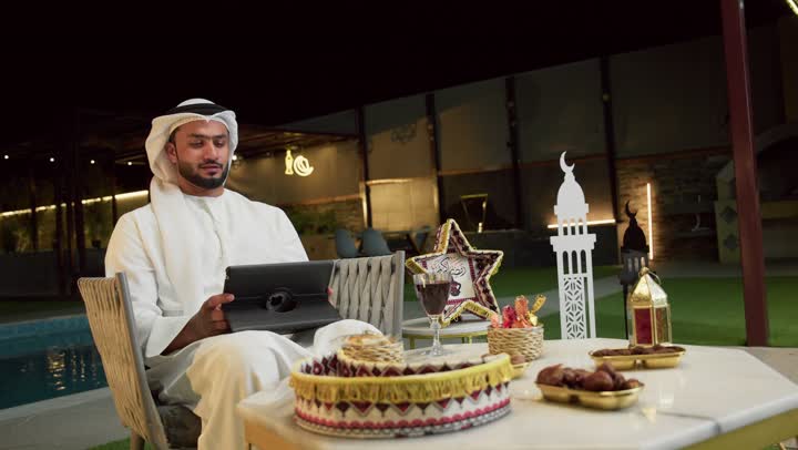 العمل عن بعد عبر الانترنت، جلسة ليلية خارجية في احدى شاليهات الامارات، ضيافة شهر رمضان المبارك، شاب عربي خليجي اماراتي يرتدي الكندورة والغترة يجلس في الهواء الطلق ويعمل على الجهاز اللوحي، استخدام جهاز التابلت بتعابير الاندماج والسرور، زينة واضاءة رمضانية