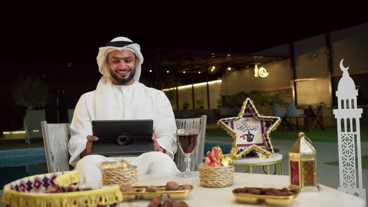 إجراء اجتماع عمل عن بعد عبر الانترنت، جلسة ليلية خارجية في احدى شاليهات الامارات، ضيافة شهر رمضان المبارك، شاب عربي خليجي اماراتي يرتدي الكندورة والغترة يجلس في الهواء الطلق ويقوم باستخدام جهاز التابلت بتعابير الاندماج والسرور، زينة واضاءة رمضانية