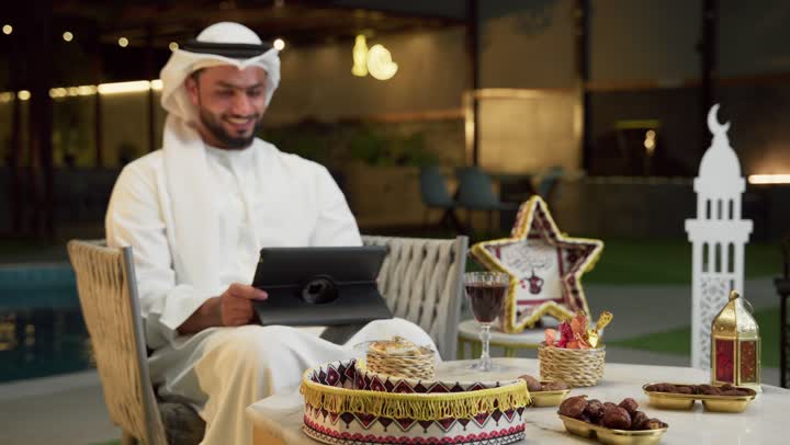 Working remotely via the internet, an outdoor night session in one of the chalets in the Emirates, hospitality during the holy month of Ramadan, a young Emirati Gulf Arab man wearing a kandura and ghutra sitting outdoors and working on a tablet, using the tablet with expressions of integration and pleasure, Ramadan decorations and lighting