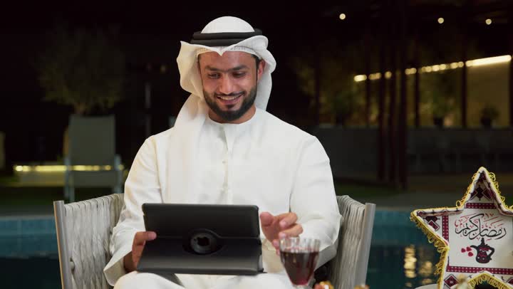 إجراء اجتماع عمل عن بعد عبر الانترنت، جلسة ليلية خارجية في احدى شاليهات الامارات، ضيافة شهر رمضان المبارك، شاب عربي خليجي اماراتي يرتدي الكندورة والغترة يجلس في الهواء الطلق ويقوم باستخدام جهاز التابلت بتعابير الاندماج والسرور، زينة واضاءة رمضانية