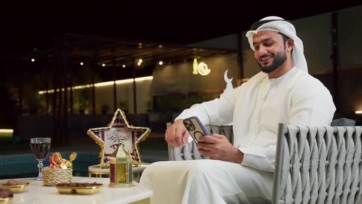استخدام جهاز تقني حديث، زينة وديكورات رمضانية، تصفح مواقع التواصل الاجتماعي، جلسة ليلية خارجية في شاليه سياحي، ضيافة شهر رمضان المبارك، شاب عربي خليجي اماراتي يرتدي الكندورة والغترة يجلس في الهواء الطلق ويستخدم الهاتف المحمول بتعابير الاندماج والسرور
