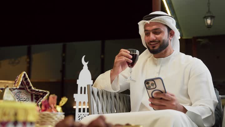 Browsing social media, an outdoor night session in a tourist chalet, Ramadan hospitality, using a modern technical device, a young Emirati Gulf Arab man wearing a kandura and ghutra sitting outdoors and using a mobile phone with expressions of integration and pleasure, Ramadan decorations and ornaments