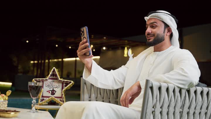 An outdoor night session in a tourist chalet, hospitality during the holy month of Ramadan, communicating with family and friends with expressions of pleasure, using a modern technical device, a young Emirati Gulf Arab man wearing a kandura and ghutra sitting outdoors and making a video call via mobile phone, Ramadan decorations and ornaments