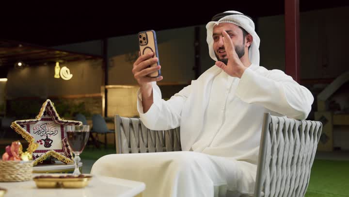ضيافة شهر رمضان المبارك، التواصل مع الأهل والأصدقاء بتعابير السرور، استخدام جهاز تقني حديث، شاب عربي خليجي اماراتي يرتدي الكندورة والغترة يجلس في الهواء الطلق ويجري مكالمة فيديو عبر الهاتف المحمول، زينة وديكورات رمضانية، جلسة ليلية خارجية في شاليه سياحي