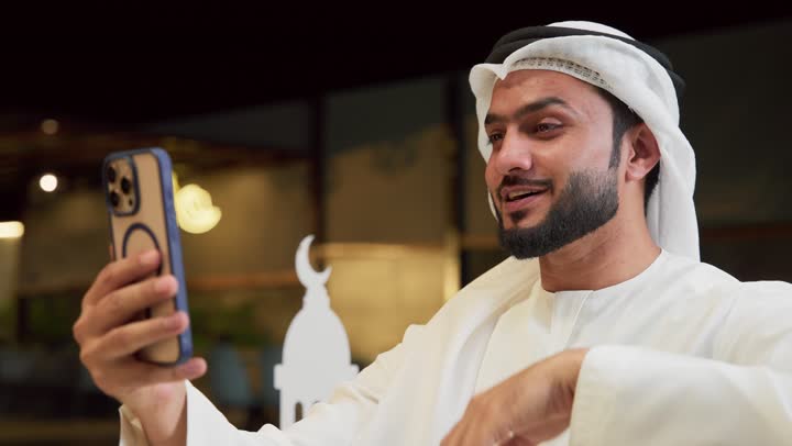 جلسة ليلية خارجية في شاليه سياحي، ضيافة شهر رمضان المبارك، التواصل مع الأهل والأصدقاء بتعابير السرور، استخدام جهاز تقني حديث، شاب عربي خليجي اماراتي يرتدي الكندورة والغترة يجلس في الهواء الطلق ويجري مكالمة فيديو عبر الهاتف المحمول، زينة وديكورات رمضانية