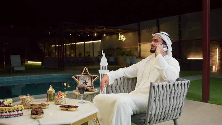 الرد على الاتصال الهاتفي بتعابير السرور، ضيافة شهر رمضان المبارك، استخدام جهاز تقني حديث، شاب عربي خليجي اماراتي يرتدي الكندورة والغترة يجلس في الهواء الطلق ويجري مكالمة عبر الهاتف المحمول، زينة وديكورات رمضانية، جلسة ليلية خارجية في احدى الشاليهات