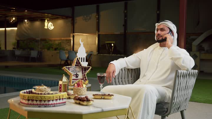 ضيافة شهر رمضان المبارك، الرد على الاتصال الهاتفي بتعابير السرور، استخدام جهاز تقني حديث، شاب عربي خليجي اماراتي يرتدي الكندورة والغترة يجلس في الهواء الطلق ويجري مكالمة عبر الهاتف المحمول، زينة وديكورات رمضانية، جلسة ليلية خارجية في احدى الشاليهات
