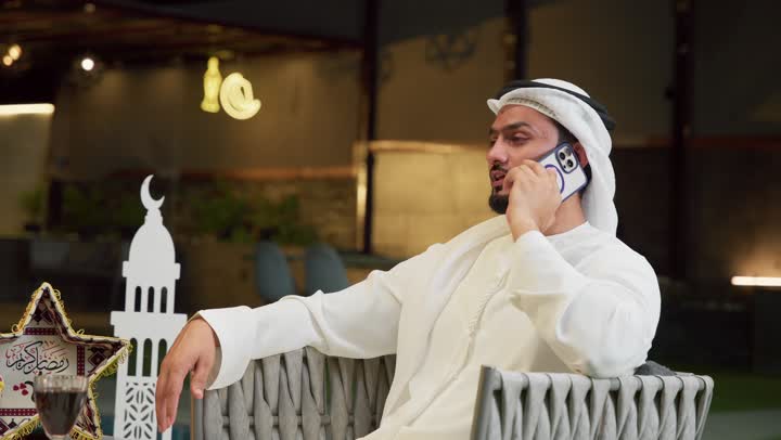Answering phone calls with happy expressions, Ramadan hospitality, using a modern technical device, a young Emirati Gulf Arab man wearing a kandura and ghutra sitting outdoors making a mobile phone call, Ramadan decorations, an outdoor night session in a chalet