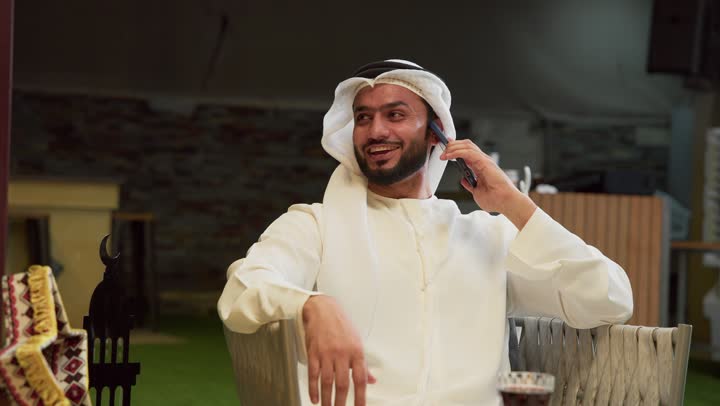 الرد على الاتصال الهاتفي بتعابير السرور، ضيافة شهر رمضان المبارك، استخدام جهاز تقني حديث، شاب عربي خليجي اماراتي يرتدي الكندورة والغترة يجلس في الهواء الطلق ويجري مكالمة عبر الهاتف المحمول، زينة وديكورات رمضانية، جلسة ليلية خارجية في احدى الشاليهات