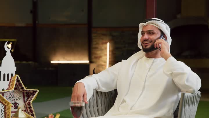 ضيافة شهر رمضان المبارك، الرد على الاتصال الهاتفي بتعابير السرور، استخدام جهاز تقني حديث، شاب عربي خليجي اماراتي يرتدي الكندورة والغترة يجلس في الهواء الطلق ويجري مكالمة عبر الهاتف المحمول، زينة وديكورات رمضانية، جلسة ليلية خارجية في احدى الشاليهات