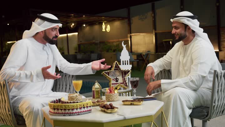 Spending a pleasant time with friends outdoors, Ramadan decorations, warm welcome and hospitality in the holy month of Ramadan, two Emirati Gulf Arab friends wearing kandura and ghutra sitting in a tourist chalet exchanging pleasant conversations, enjoying cold drinks