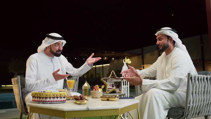 قضاء وقت ممتع مع الاصدقاء في الهواء الطلق، زينة وديكور رمضاني، حسن الاستقبال وكرم الضيافة في شهر رمضان المبارك، صديقان عربيان خليجيان اماراتيان يرتديان الكندورة والغترة يجلسان في شاليه سياحي يقومان بتبادل الاحاديث بتعابير السرور، الاستمتاع بتناول المشروبات الباردة