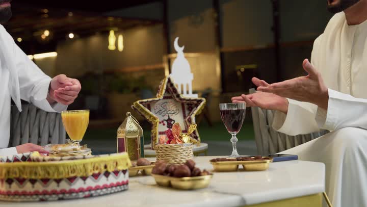 A table full of Ramadan decorations and ornaments, warm welcome and hospitality during the holy month of Ramadan, two Emirati Gulf Arab friends wearing kanduras and ghutras sitting in a tourist chalet exchanging conversations with happy expressions, enjoying cold drinks