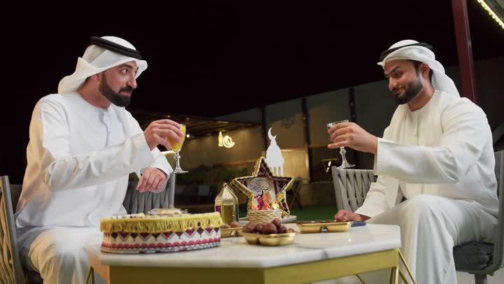 قضاء وقت ممتع مع الاصدقاء في الهواء الطلق، زينة وديكور رمضاني، حسن الاستقبال وكرم الضيافة في شهر رمضان المبارك، صديقان عربيان خليجيان اماراتيان يرتديان الكندورة والغترة يجلسان في شاليه سياحي يقومان بتبادل الاحاديث بتعابير السرور، الاستمتاع بتناول المشروبات الباردة