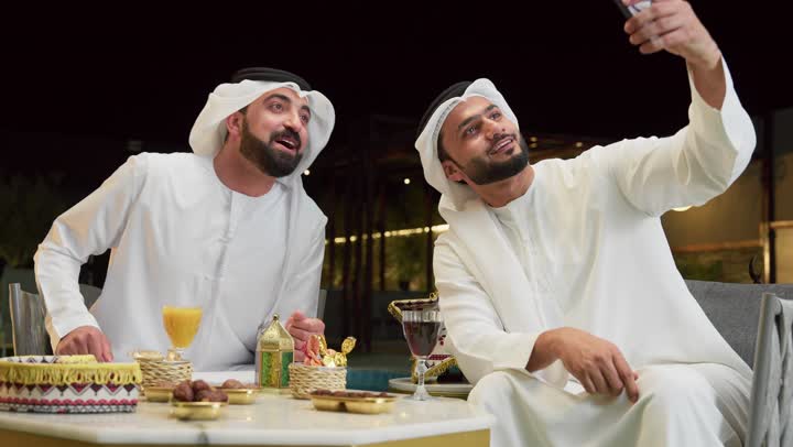 Documenting happy moments, using the mobile phone to express joy and happiness, Ramadan decorations, warm welcome and hospitality during the holy month of Ramadan, two Emirati Gulf Arab friends wearing kandura and ghutra sitting in a tourist chalet, taking a selfie using a mobile phone