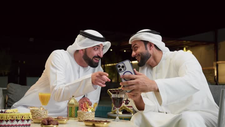 قضاء وقت ممتع مع الأصدقاء، استخدام الهاتف المحمول بتعابير السرور والسعادة، زينة وديكور رمضاني، حسن الاستقبال وكرم الضيافة في شهر رمضان المبارك، رجلان عربيان خليجيان اماراتيان يرتديان الكندورة والغترة يجلسان في شاليه سياحي، تصفح مواقع الانترنت
